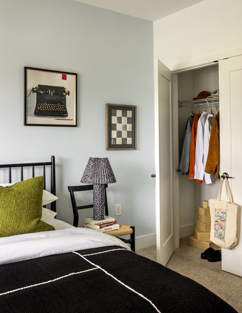 A bedroom with a bed, a chair, a lamp, a picture of a typewriter, a picture of a checkerboard, a wardrobe with clothes, and a tote bag on the floor.