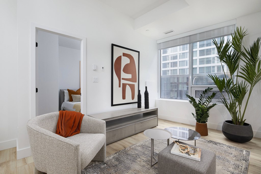 A living room with a grey armchair, a grey coffee table, a large framed artwork on the wall, and a large window with a view of a cityscape.