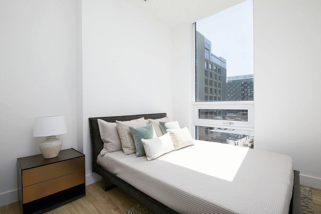 A bedroom with a large bed, a nightstand with a lamp, and a window with a view of a cityscape.
