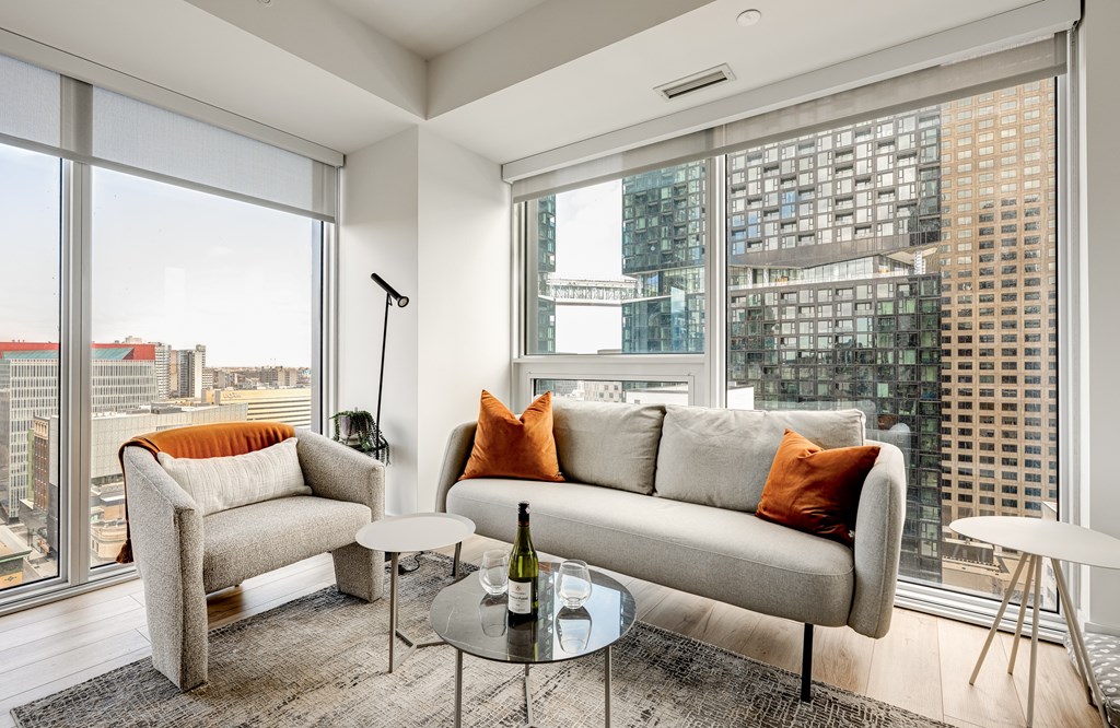 A living room with a grey couch, a glass table, and a large window with a view of the city.