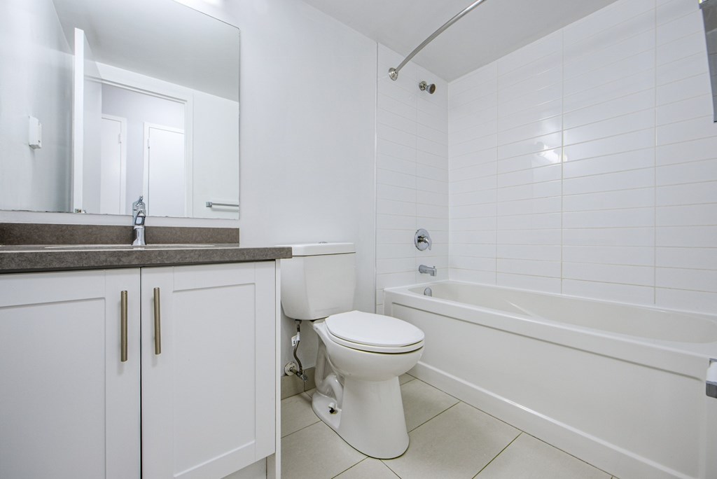 A white toilet sits in a bathroom with a white tub and a mirror.
