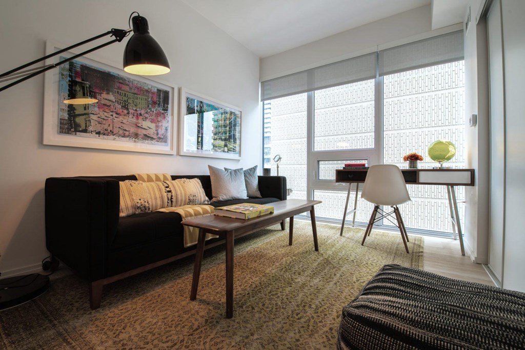 A living room with a black couch, a table with a lamp, a chair, and a window with blinds.