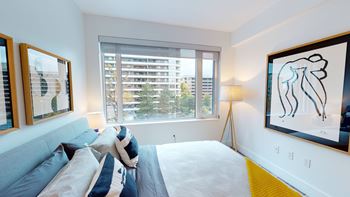 A bedroom with a bed, a window with blinds, and two framed pictures on the wall.
