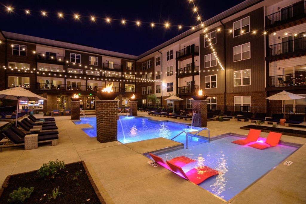 A pool area with a lit up water feature and lounge chairs.