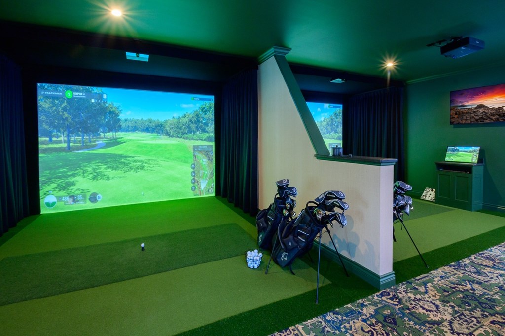 A golf simulator room with a screen, golf clubs, and a golf ball on the green carpet.