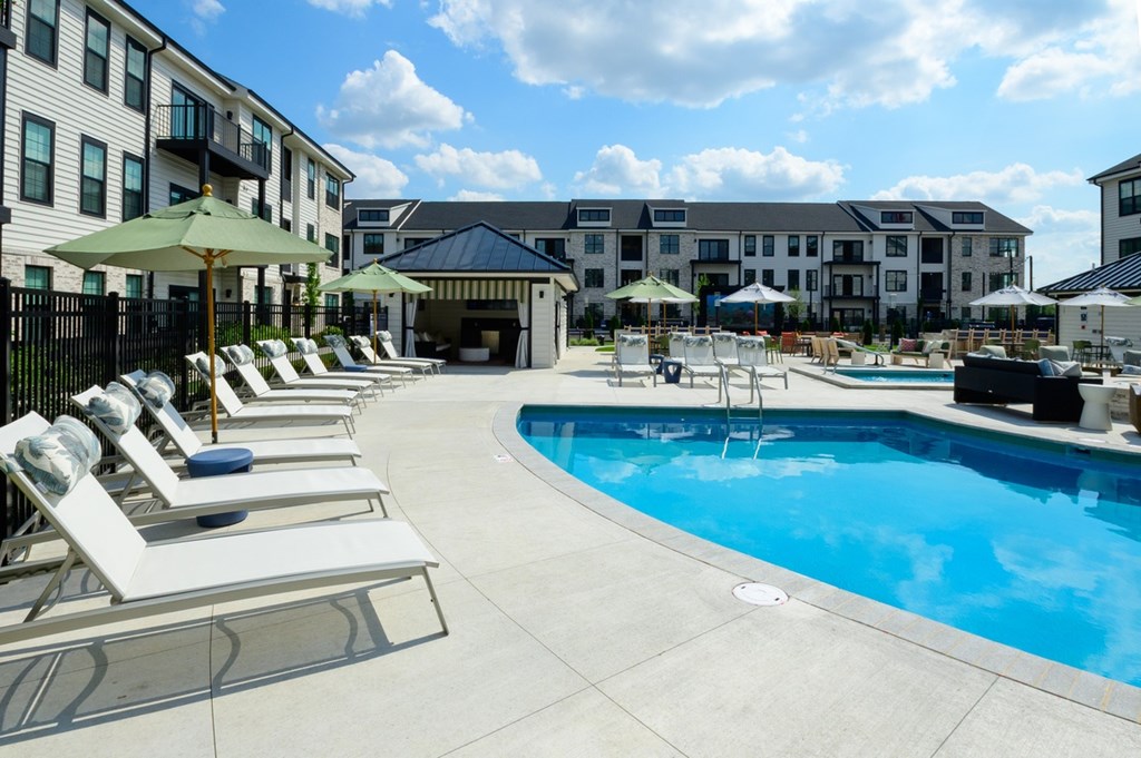 A swimming pool surrounded by sun loungers and umbrellas in front of apartment buildings.