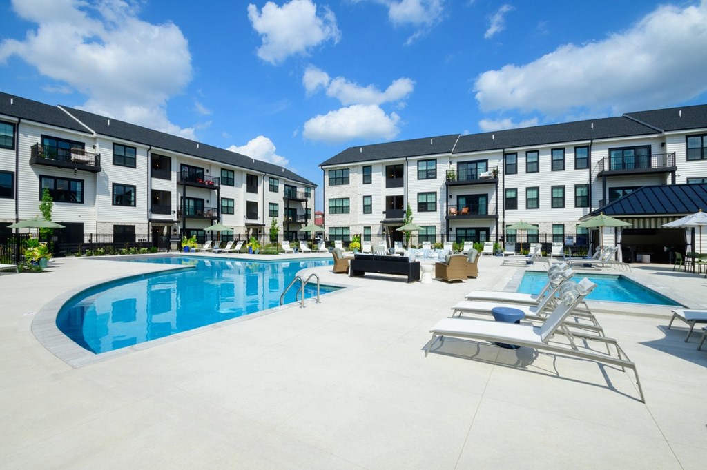 A large swimming pool is surrounded by lounge chairs in front of apartment buildings.