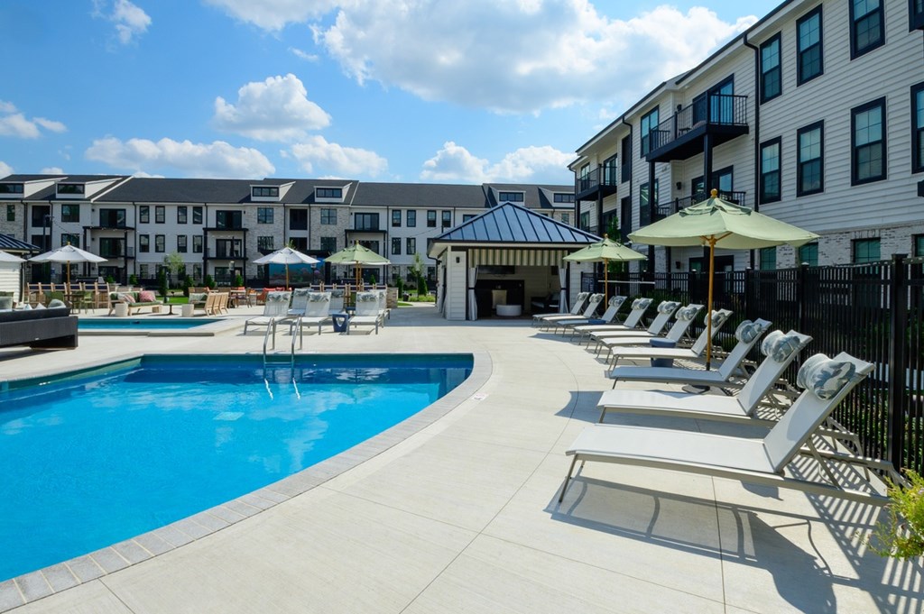 A swimming pool surrounded by sun loungers and umbrellas in front of apartment buildings.