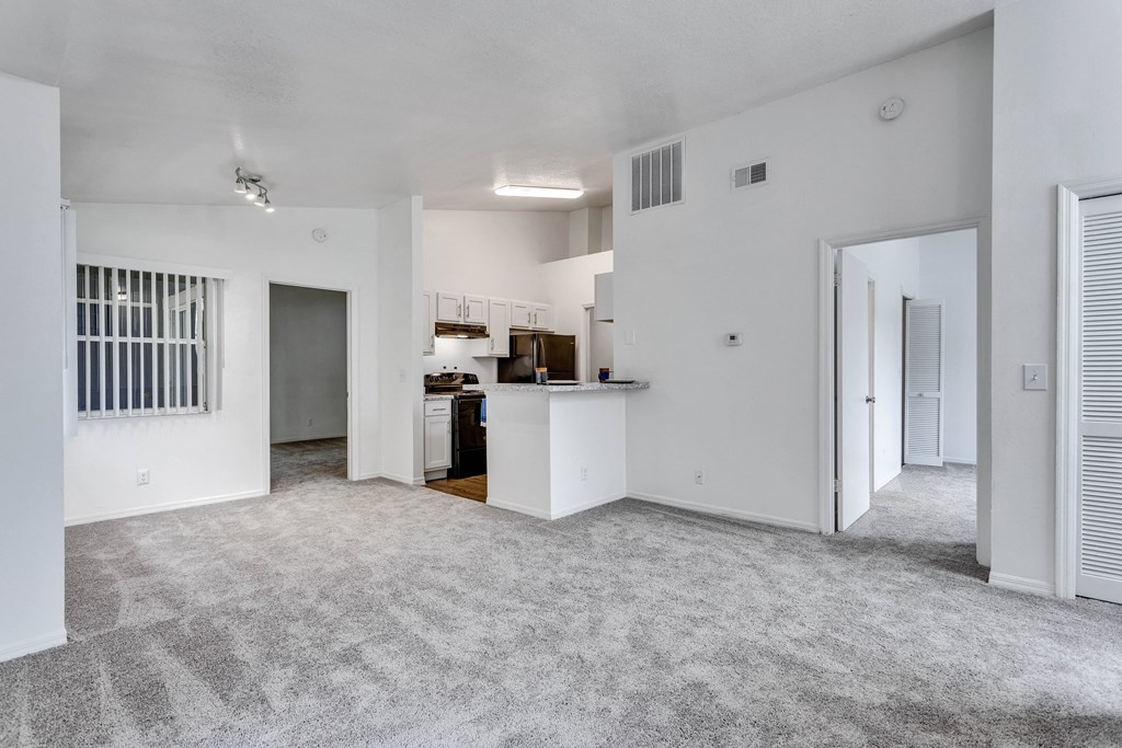 the living room and kitchen of an apartment with white walls and carpet