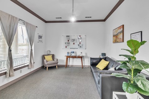 A living room with a grey couch, a yellow pillow, a grey armchair, a wooden side table, a white wall with pictures and a plant.