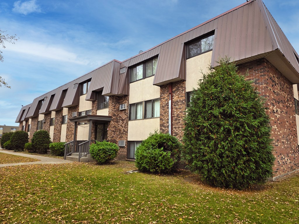 A building with brown and beige exterior and a brown roof with windows and bushes in front.