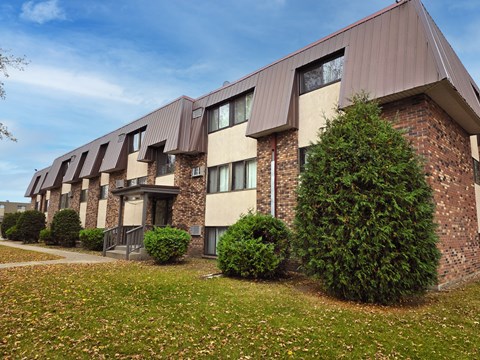 A building with brown and beige exterior and a brown roof with windows and bushes in front.