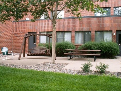 A tree stands in front of a brick building with a swing set and two benches.