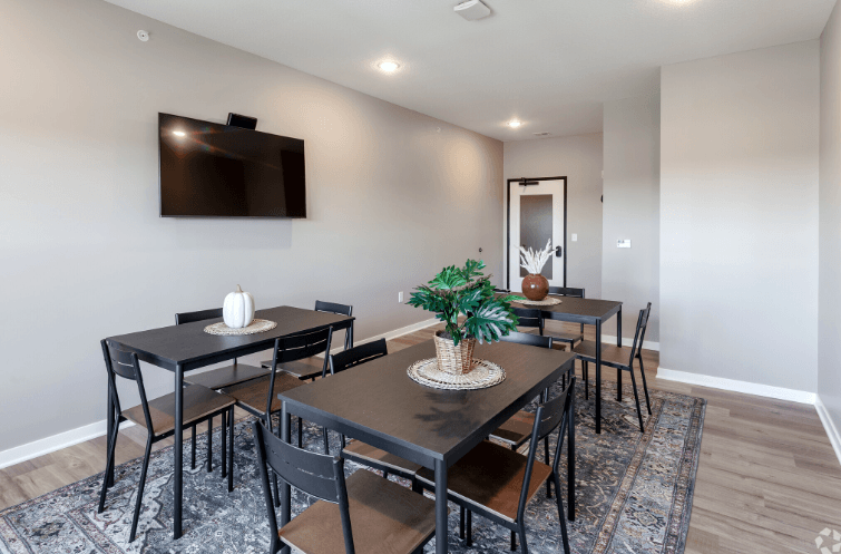 A black table with a vase and a basket on it is surrounded by chairs in a room with a flat screen TV mounted on the wall.