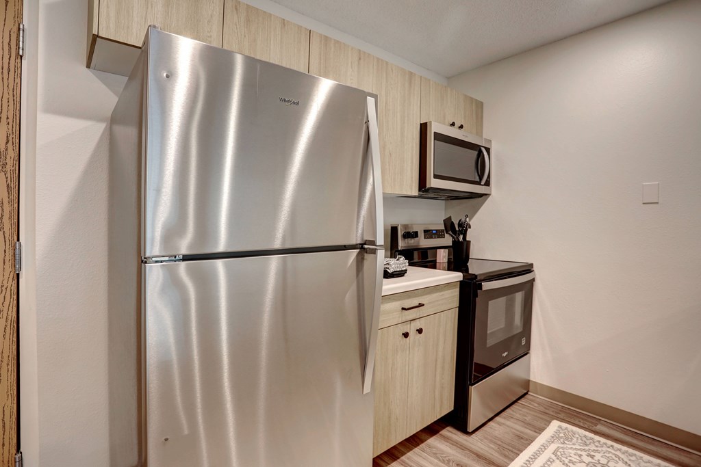 A stainless steel refrigerator stands in a kitchen with wooden cabinets and a microwave above the stove.