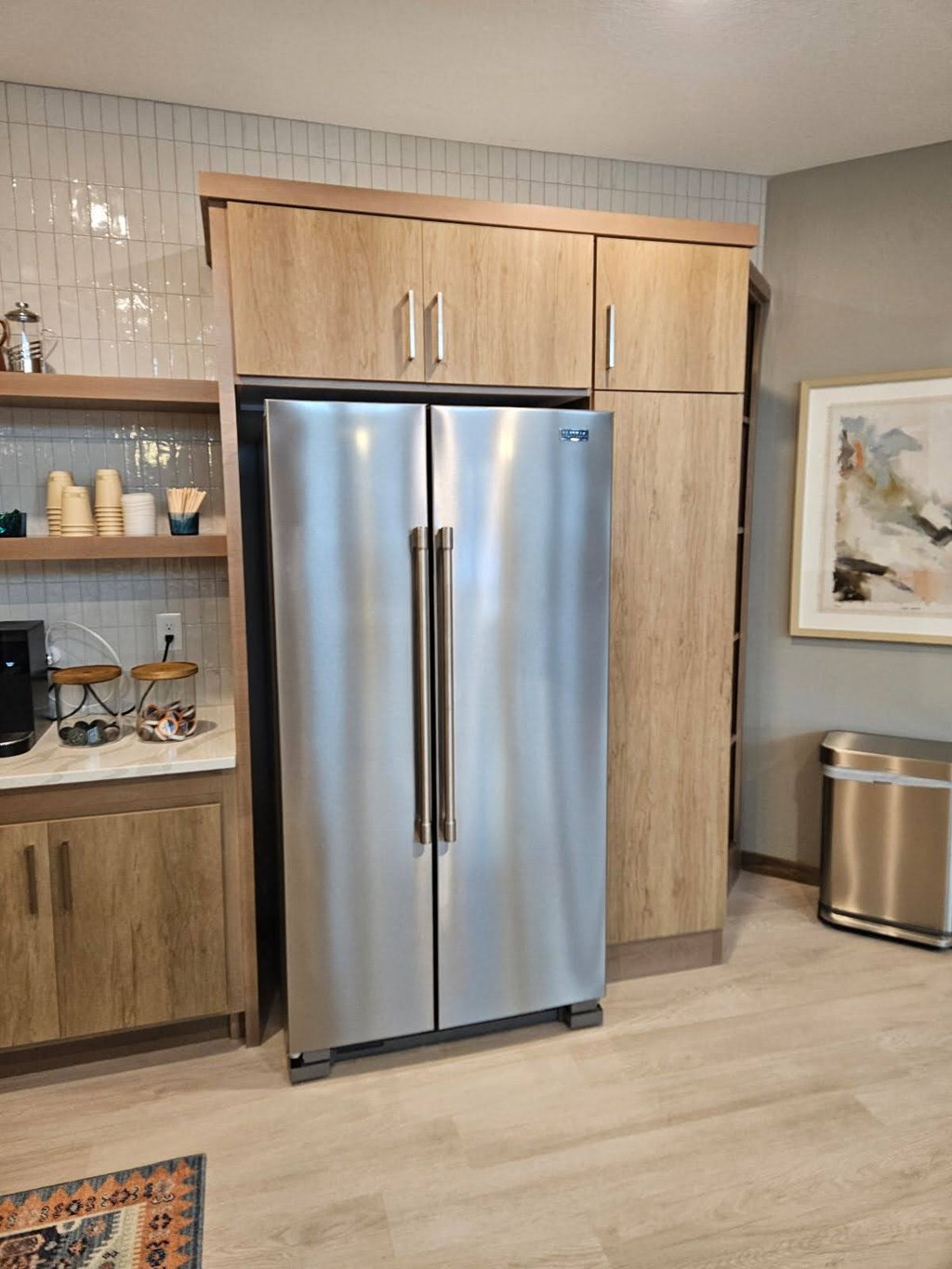 A stainless steel refrigerator with wooden cabinets and a trash can in a kitchen.