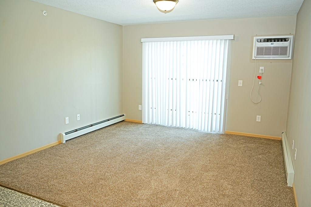 A room with a carpeted floor, a window with blinds, and a wall-mounted air conditioner unit.