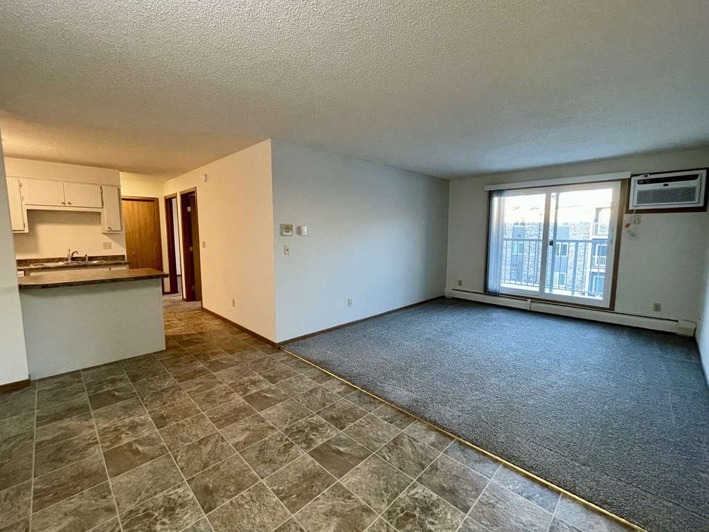 A spacious living room with a kitchen area to the left and a window with a view of a building outside.