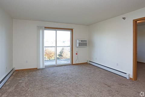 A room with a carpet floor, a window, a door, and a wall-mounted air conditioner unit.