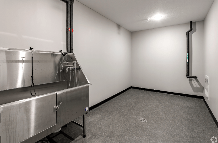 A large metal stainless steel industrial tank with a hose connected to it is in a room with grey carpeting.