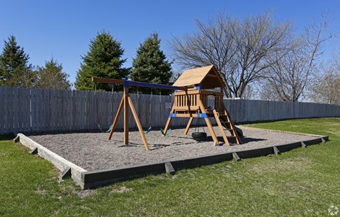 A wooden swing set with a brown roof is situated on a gravel area in a backyard.