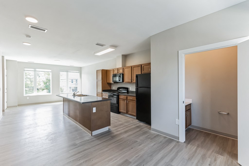 an empty living room with a kitchen with a black refrigerator and