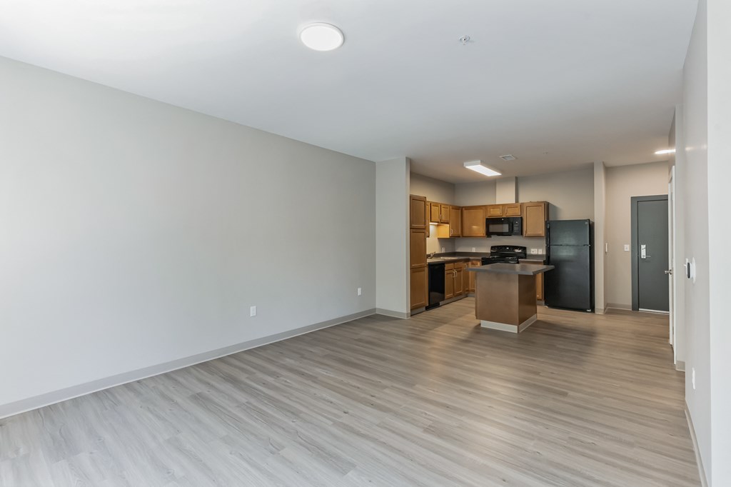 an empty living room with a kitchen with a black refrigerator and
