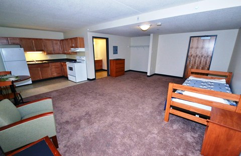 A bedroom with a bed, chair, and a kitchen area with a refrigerator, microwave, and oven.