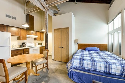 A bedroom with a bed, a table and chairs, and a kitchen area with a refrigerator, microwave, and cabinets.
