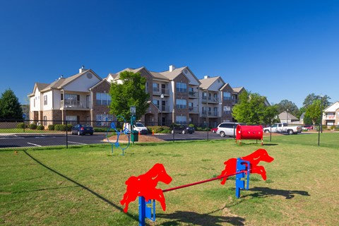 A red horse-shaped swing set sits in a grassy field in front of apartment buildings.