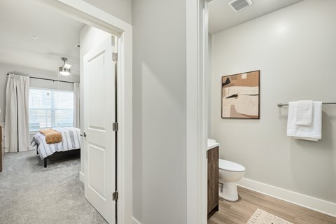 A white bathroom with a toilet, sink, and a bedroom with a bed and window.