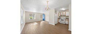 A well-lit, empty room with a chandelier and a kitchenette in the background.