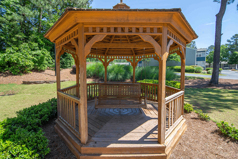 A wooden gazebo with a circular stone base is surrounded by greenery.
