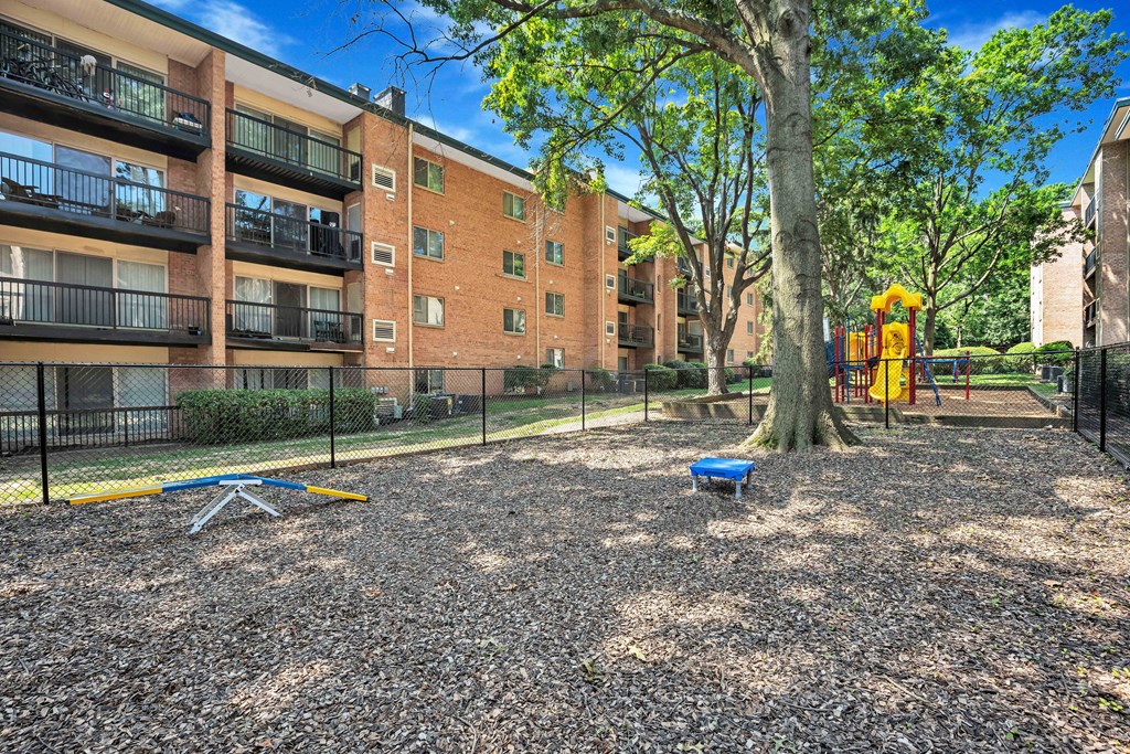 A playground area with a slide, swings, and a tree in front of a brick apartment building.