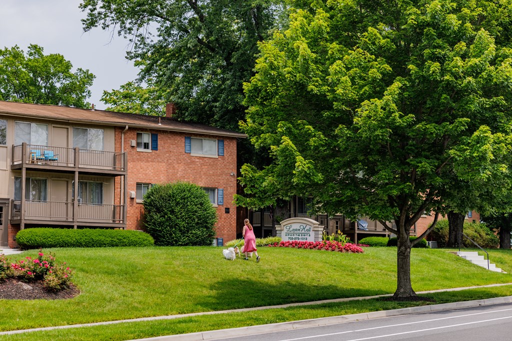 A woman in a pink dress is sitting on a lawn in front of a brick apartment building.