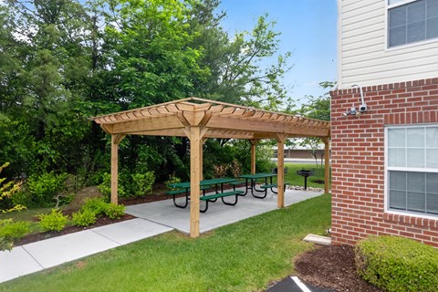 A wooden pergola with green benches is in front of a brick building.