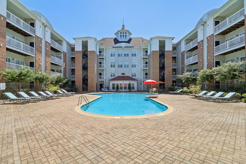 A large swimming pool surrounded by a brick patio and lounge chairs in front of apartment buildings.