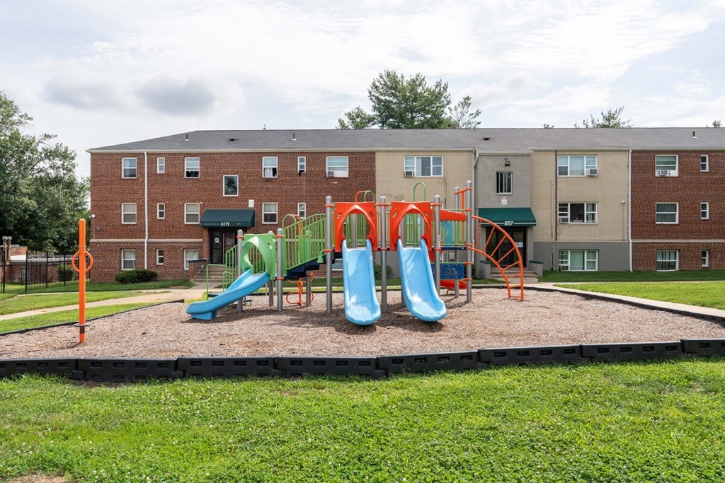 A playground with a green slide and orange safety barriers in front of a brick building.
