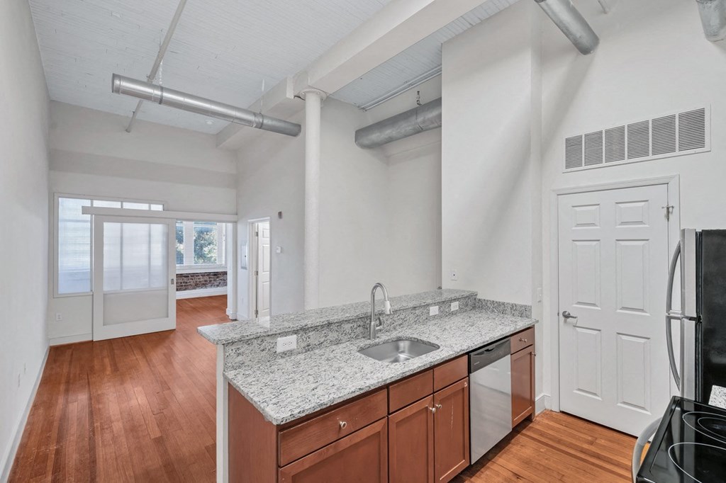 an apartment kitchen is shown with original wood floors, and granite countertop. Exposed pipes and sliding door to bedroom show character.