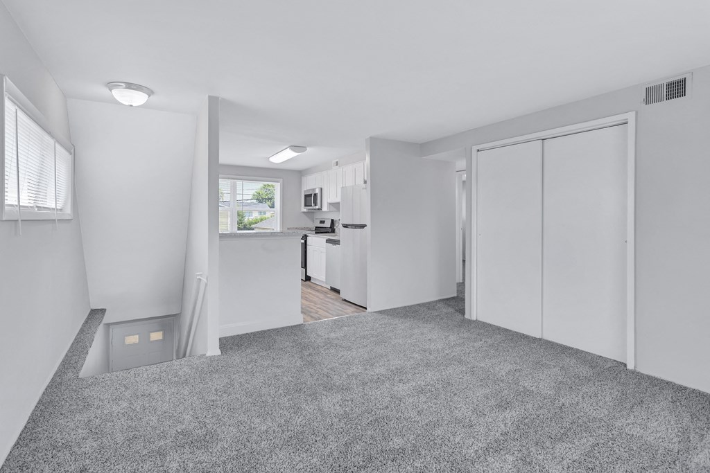 the living room and kitchen of an apartment with white walls and carpet