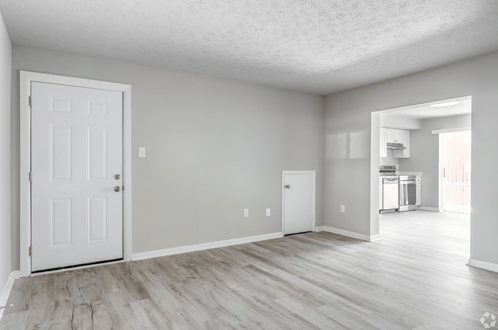 an empty living room with a white door and a kitchen