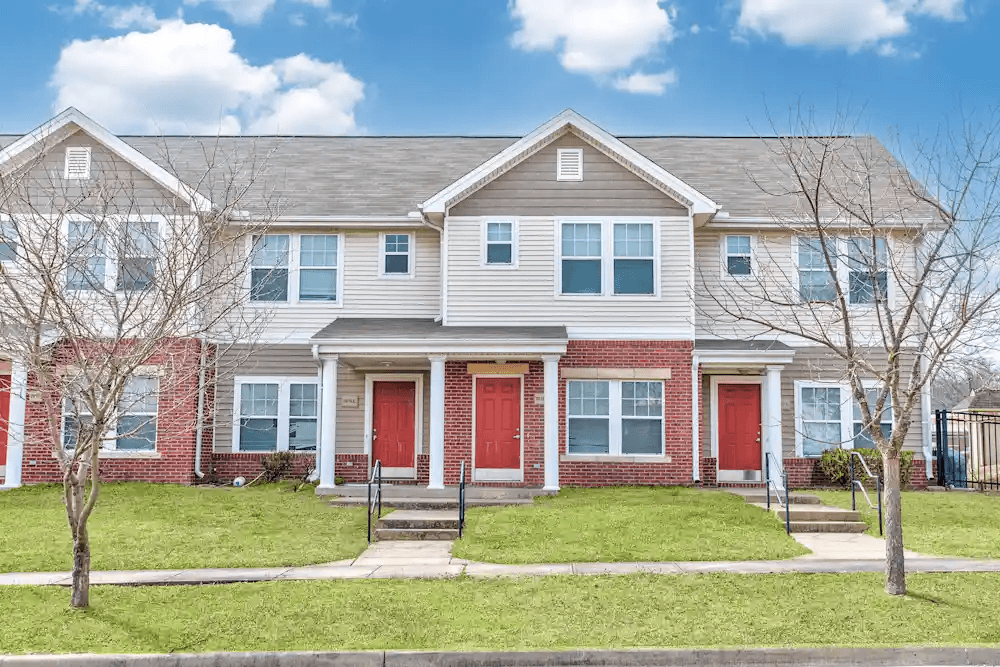 A house with a red front door is surrounded by a green lawn and leafless trees.