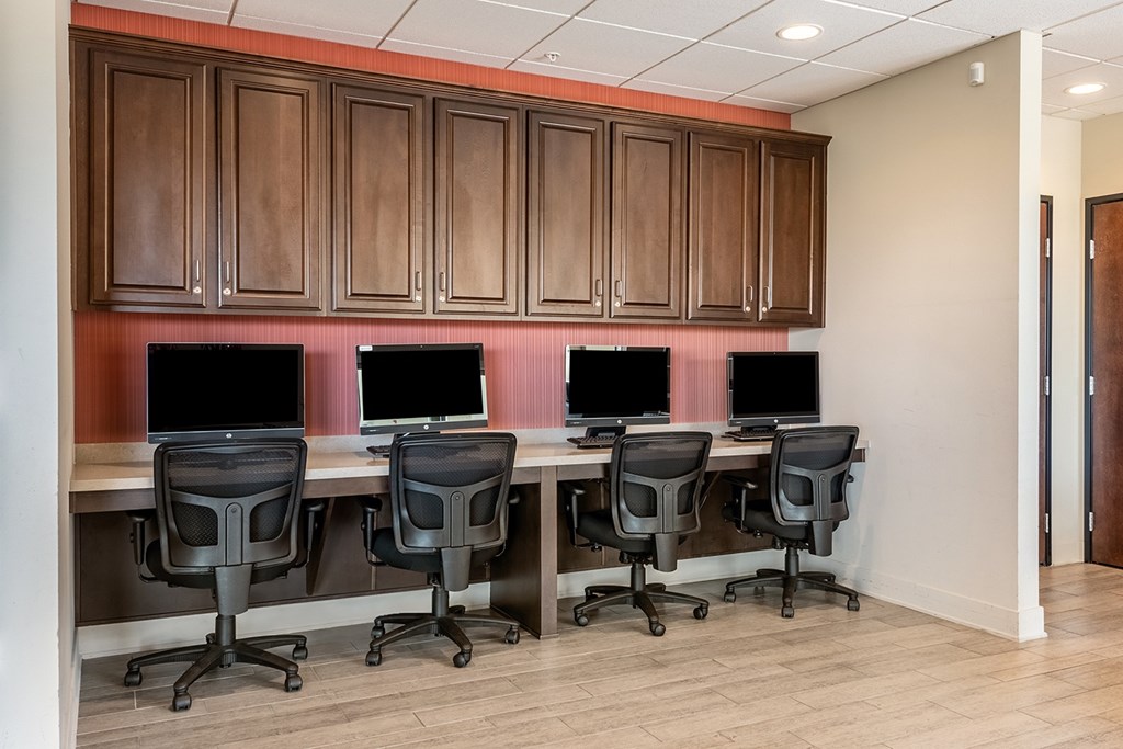 A row of four computers are on a desk with black chairs.