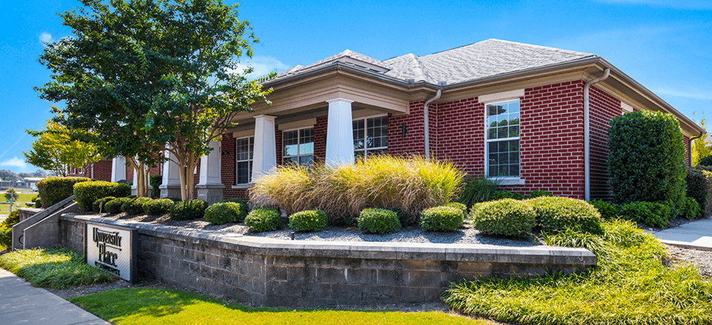 the front of a brick leasing office with a stone retaining wall and landscaping at University Place Apartments