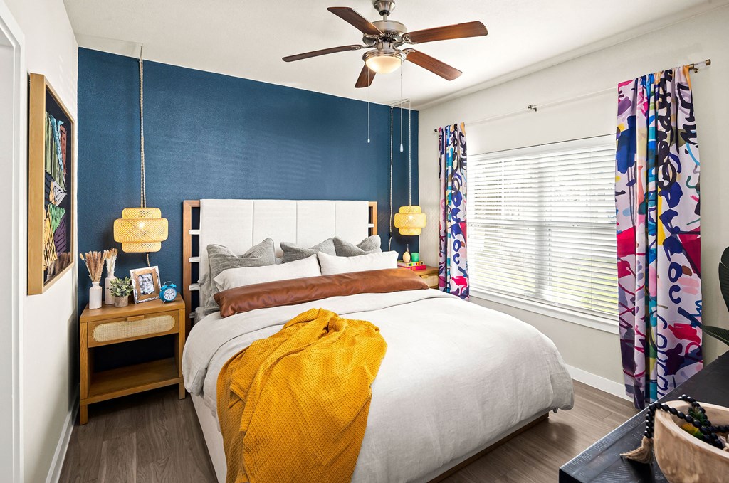 bedroom with blue accent wall behind bed. ceiling fan with handing accent lighting. bright curtains and moody decor.