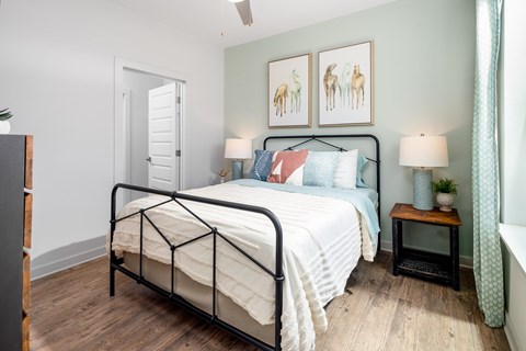 A bedroom with a black metal frame bed, a white comforter, and a small wooden table with a lamp on it.