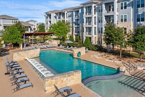 A swimming pool surrounded by sun loungers in front of apartment buildings.
