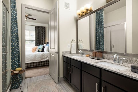 A bathroom with a mirror, sink, and a bedroom with a bed and a window.