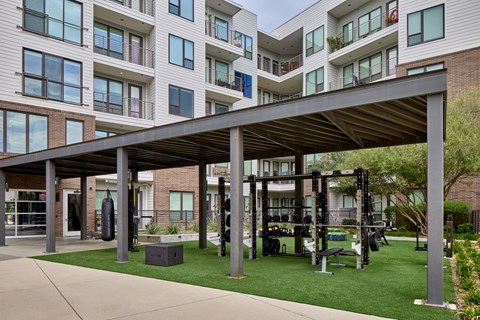 A gym area with a roofed structure and weights is located in front of apartment buildings.