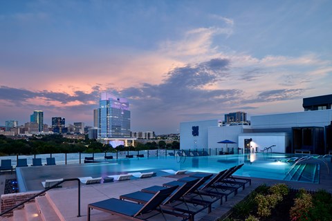 A rooftop pool area with lounge chairs and a city skyline at dusk.